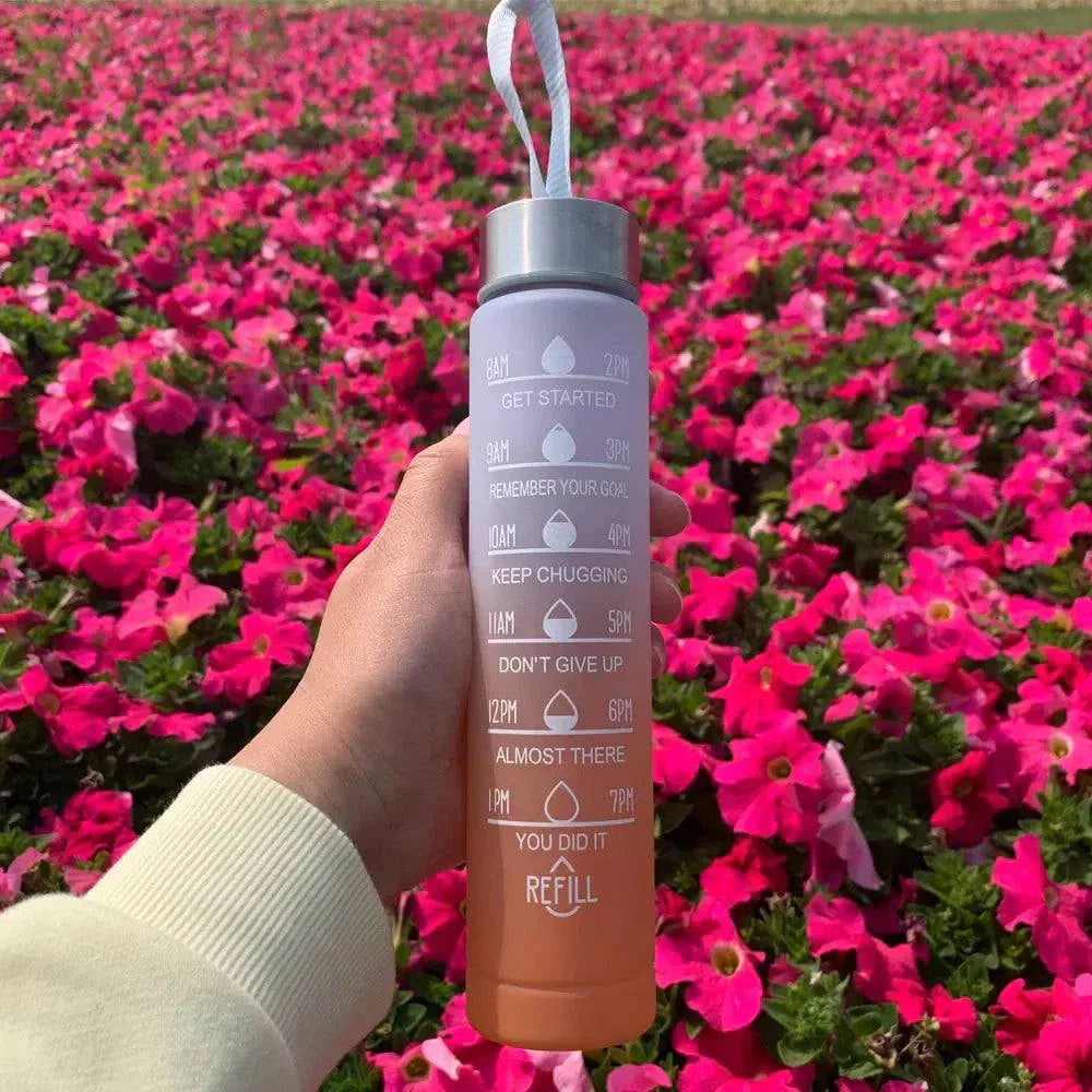 Hand holding a purple and orange water bottle with motivational quotes and time markers against a background of vibrant pink flowers.