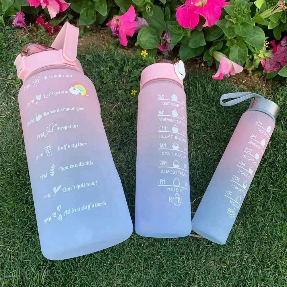 Set of three reusable plastic water bottles with motivational quotes and time markers in pink shade placed on grass with pink flowers in background