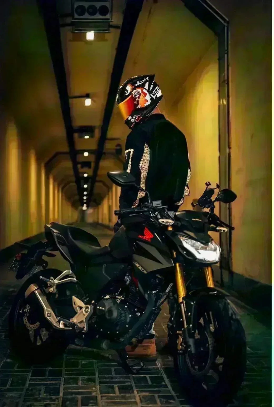 Motorcyclist in black gear and helmet standing next to a black sports motorcycle in a dimly lit tunnel