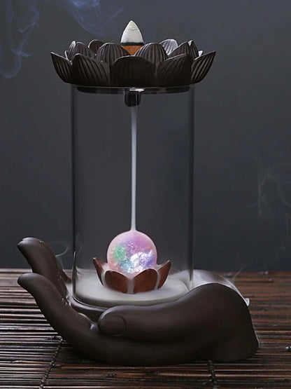 Backflow Purple Sand incense burner with glass lotus and Buddha's hand design releasing smoke cascade in ceramic hand-shaped base.