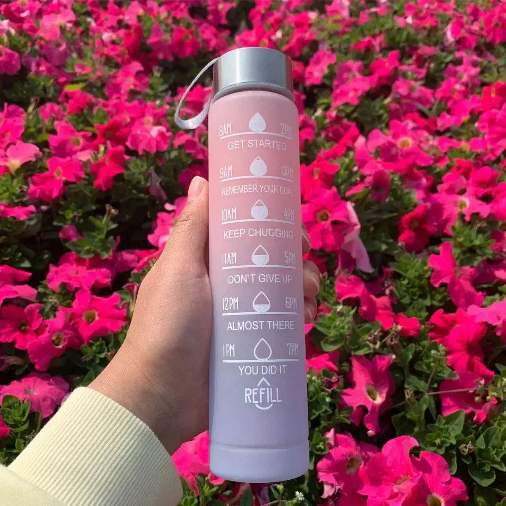Pink sports water bottle with motivational quotes and time markers, held against bright pink flowers background