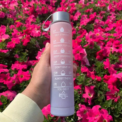 Pink sports water bottle with motivational quotes and time markers, held against bright pink flowers background