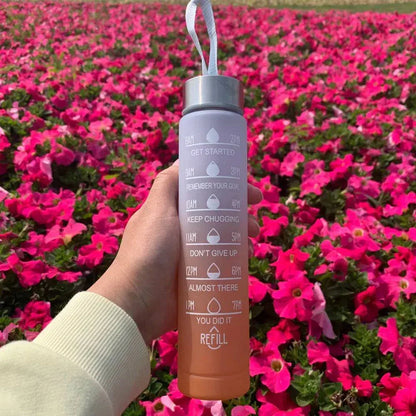 Hand holding a purple and orange water bottle with motivational quotes and time markers against a background of vibrant pink flowers.