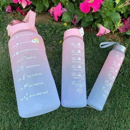 Set of three reusable plastic water bottles with motivational quotes and time markers in pink shade placed on grass with pink flowers in background